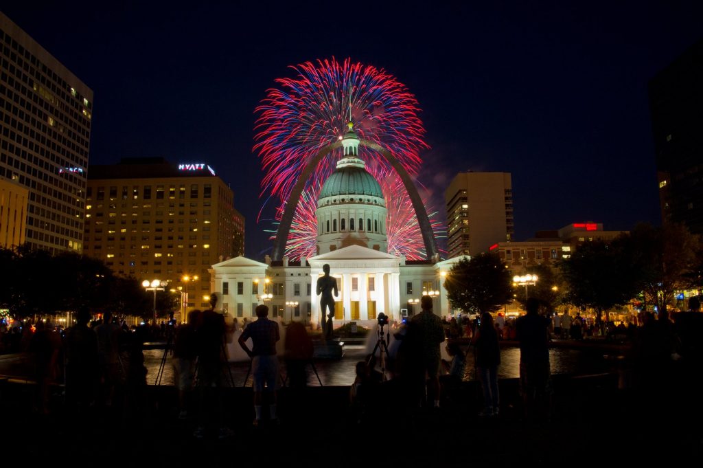 Red and blue fireworks pop against a dark night sky above the Old Courthouse and Gateway Arch in downtown St. Louis.