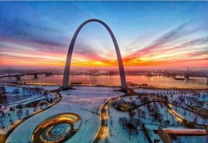Purple, pink and orange skies surround the Gateway Arch and snowy park grounds on a winter morning