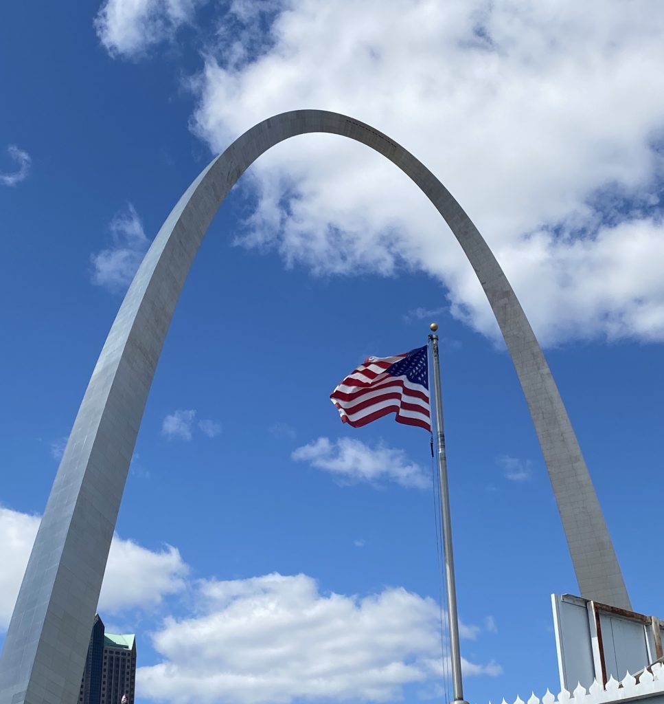 The American flag flies in the wind as the Gateway Arch stands tall behind it.