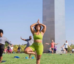 A woman stands on one leg in tree pose during a yoga session with dozens of other people under the Gateway Arch.