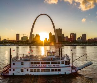 The Becky Thatcher riverboat floats past the Gateway Arch during sunset.