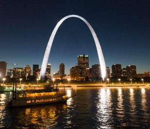 The Becky Thatcher riverboat cruises at night along the Mississippi River in front of the illuminated St. Louis Riverfront and Gateway Arch.