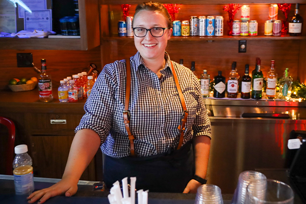 A female bartender on the Riverboats at the Gateway Arch smiles as she stands behind the bar with alcoholic drink options on the shelves behind her.