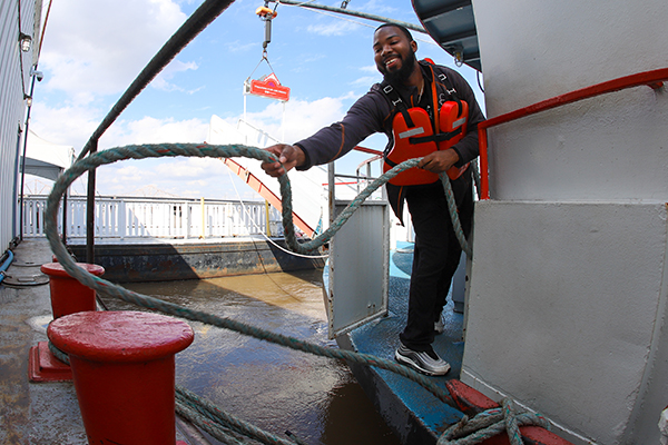 A deckhand wearing an orange life vest throws a thick hitching rope from the Riverboat around a large rung on the dock to tie up the boat for safety.