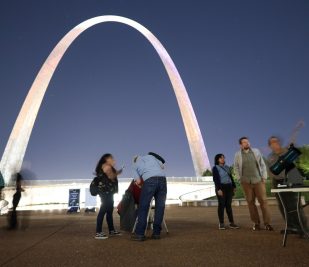 Visitors look into large telescopes sitting in front of the Gateway Arch during a Gateway to the Stars event