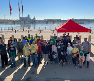 A group of about 2 dozen volunteers stand for a photo in front of the Mississippi River on the Gateway Arch National Park grounds with trash bags and pickers, ready to collect trash and clean up the St. Louis riverfront.