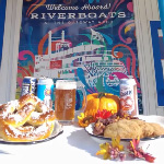 Food and drink featured on the Oktoberfest Cruise is placed on a table in front of a mural of the Riverboats at the Gateway Arch.