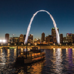 The Becky Thatcher riverboat cruises at night along the Mississippi River in front of the illuminated St. Louis Riverfront and Gateway Arch.