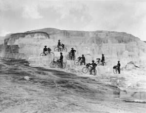 Historic black and white photo of the Iron Riders posing for a photo with their bicycles on the side of a rock formation. Image provided by National Park Service.