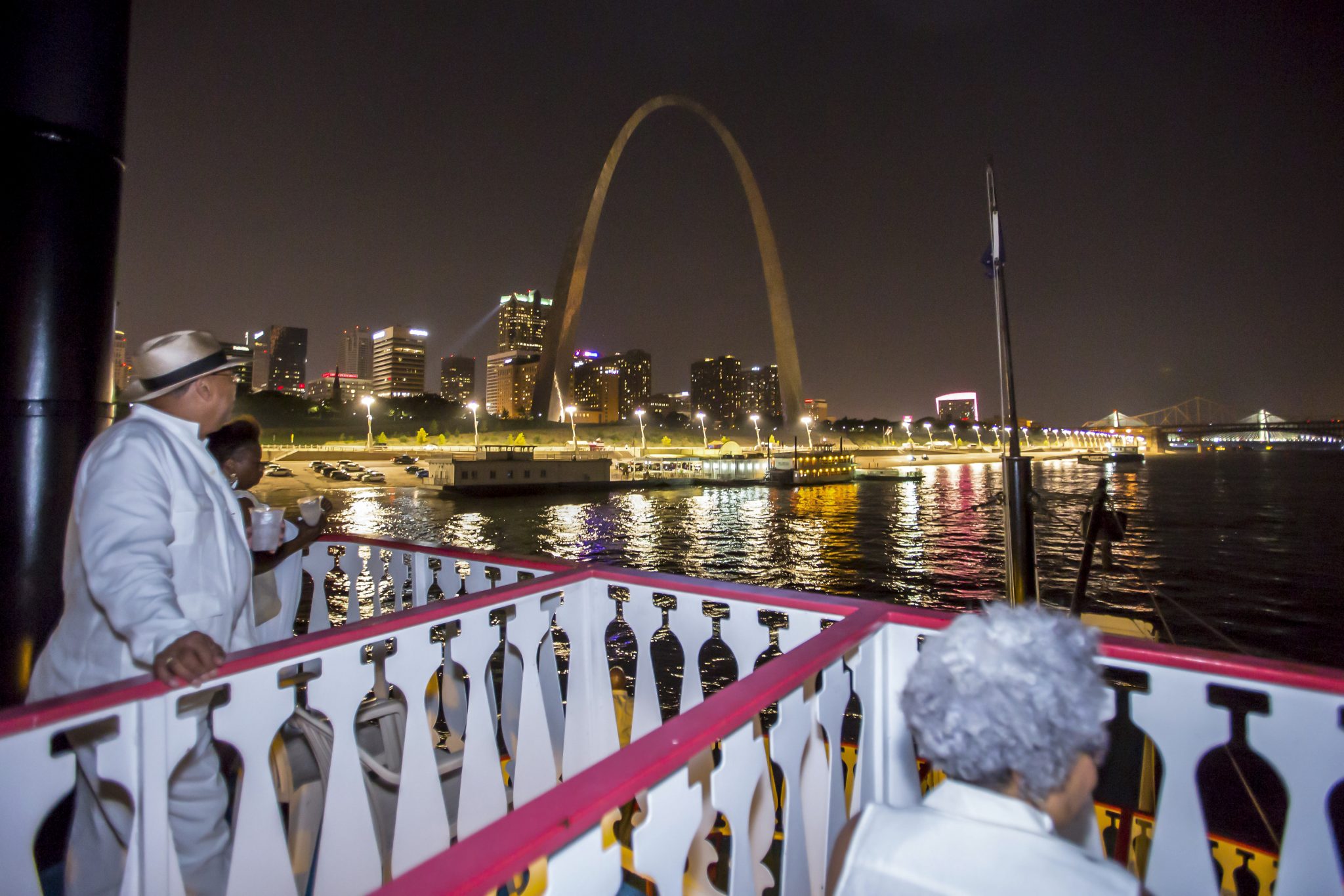 Guests sit on the top deck of the Riverboats at the Gateway Arch at night looking at the Gateway Arch illuminated