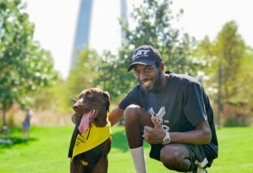 A man showing a thumbs up while crouching down next to his dog wearing a BARK ranger bandana.
