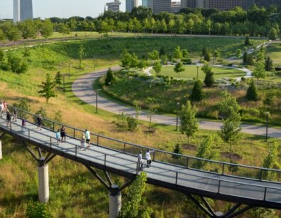 View of the North Gateway of Gateway Arch National Park. Includes the Gateway Arch, Explorer's Garden and elevated walkway with people walking on it.