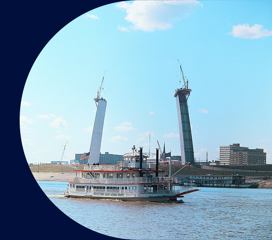 A riverboat that has "Huck Finn Sight-Seeing Tours" on the side is cruising on the Mississippi river in front of a half built arch circa 1964.