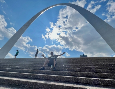 Man sitting and posing with arms wide on the Grand Staircase under the arch against a blue cloud filled sky.