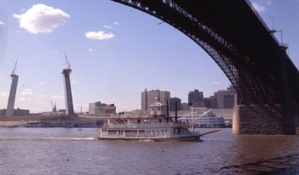 The Huck Finn Sight-Seeing Tours riverboat is cruising under the Eads Bridge with a partially finished Gateway Arch in the background circa 1964.