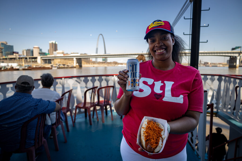 Riverboat passenger in a St. Louis shirt, holding a drink and hot dog with the Gateway Arch in the background.