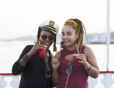 Two women holding drinks cheers and smile to the camera on the outside deck of a Groove & Spin Cruise on the Riverboats at the Gateway Arch