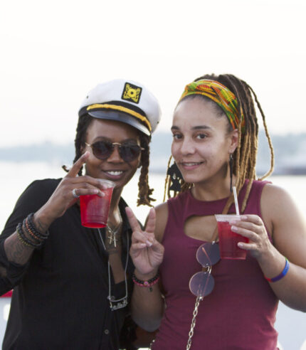 Two women holding drinks cheers and smile to the camera on the outside deck of a Groove & Spin Cruise on the Riverboats at the Gateway Arch