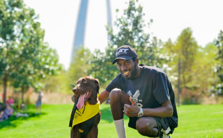 Gateway Arch visitor poses with his chocolate lab dog, wearing a B.A.R.K. Ranger bandana at Gateway Arch National Park