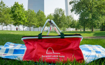 A red picnic basket sits in the grass under the Gateway Arch