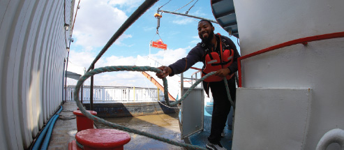 A deckhand wearing an orange life vest throws a thick hitching rope from the Riverboat around a large rung on the dock to tie up the boat for safety.