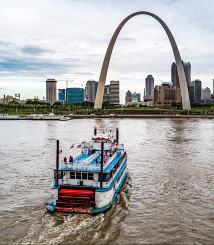 Tom Sawyer riverboat cruising on the Mississippi River with the St. Louis skyline and Gateway Arch in the background.