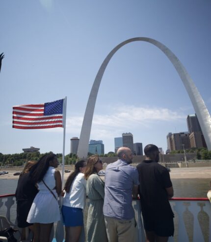 A group of people stand on the top deck of the Tom Sawyer Riverboat, looking up at the Gateway Arch during a cruise.