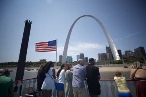 A group of people stand on the top deck of the Tom Sawyer Riverboat, looking up at the Gateway Arch during a cruise.