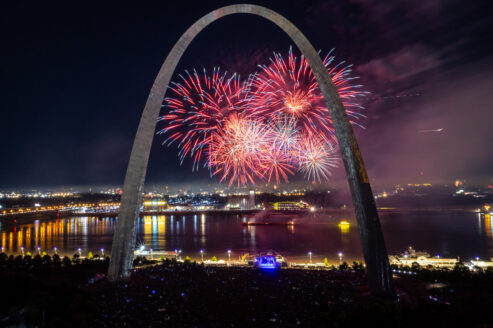 An aerial image shows a large crowd gathered under the Gateway Arch at night with 4th of July fireworks exploding over the Mississippi River.