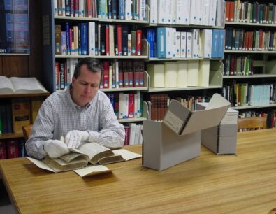 A man wearing while gloves carefully handles a book in the archives of a museum, surrounded by book shelves.