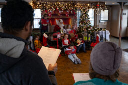 Santa and a group of children sing Christmas carols, with parents standing and singing along too.