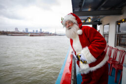 Santa rides on the Tom Sawyer riverboat, leaning over the railing to look out at downtown St. Louis.