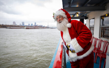 Santa rides on the Tom Sawyer riverboat, leaning over the railing to look out at downtown St. Louis.