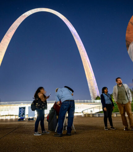 People look into telescopes in front of the Gateway Arch at night during a Gateway to the Stars event, with cutout image of Park Ranger Richard Fefferman