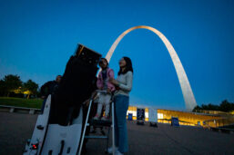 A woman and her young daughter look through a telescope into the night sky in front of the Gateway Arch