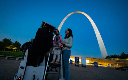 A woman and her young daughter look through a telescope into the night sky in front of the Gateway Arch