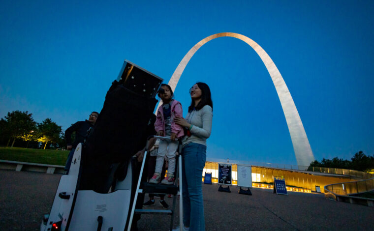 A woman and her young daughter look through a telescope into the night sky in front of the Gateway Arch