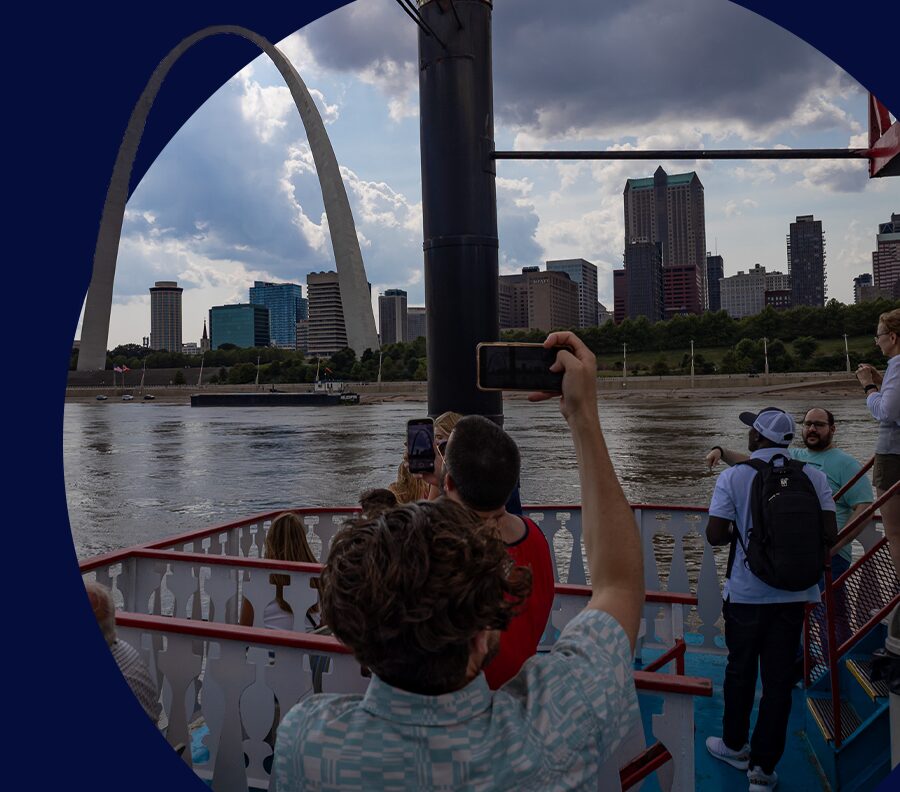 A young man takes a photo with his phone of the Gateway Arch from the top deck of the Tom Sawyer Riverboat in the Mississippi River