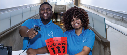 A pair of Gateway Arch Tour Guides smile in a photo as they hold up boarding passes and a ticket scanner before a Tram Ride to the Top.
