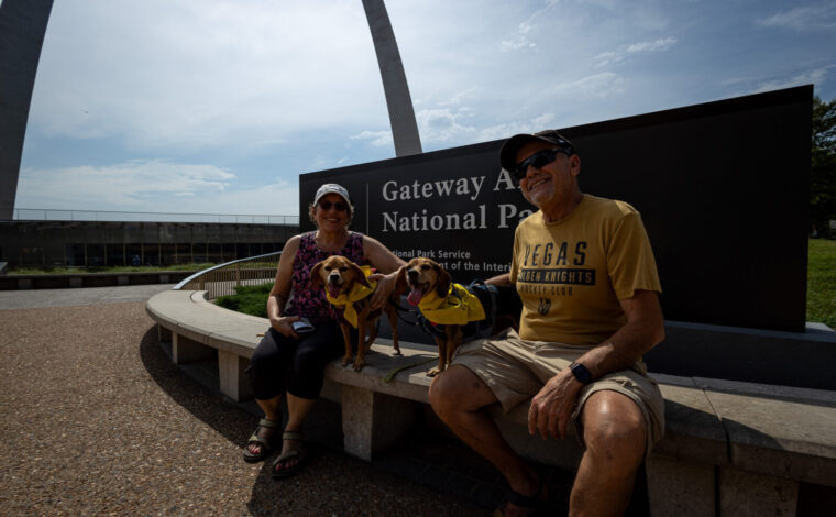 Two visitors and their dogs in BARK Ranger bandanas sit in front of the Gateway Arch National Park sign.