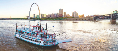 The Tom Sawyer riverboat floating in the Mississippi River with the Gateway Arch and St. Louis skyline in background.