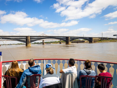 Passengers sit on the top deck of a riverboat and look over at Eads Bridge on the Misssissippi River.