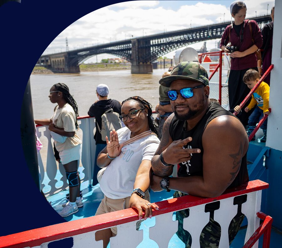 Hero graphic of guests smiling and posing for a photo on the top deck of the Tom Sawyer riverboat during a St. Louis Riverfront Cruise.