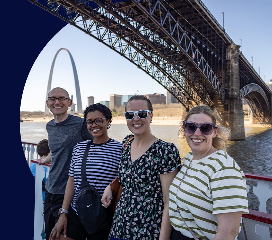 Website hero graphic of a group of four people smiles for a photo on riverfront cruise with Eads Bridge and the Arch in the background