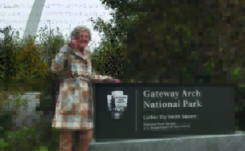 Grandma Joy stands in front of a Gateway Arch National park sign with the Gateway Arch in the background